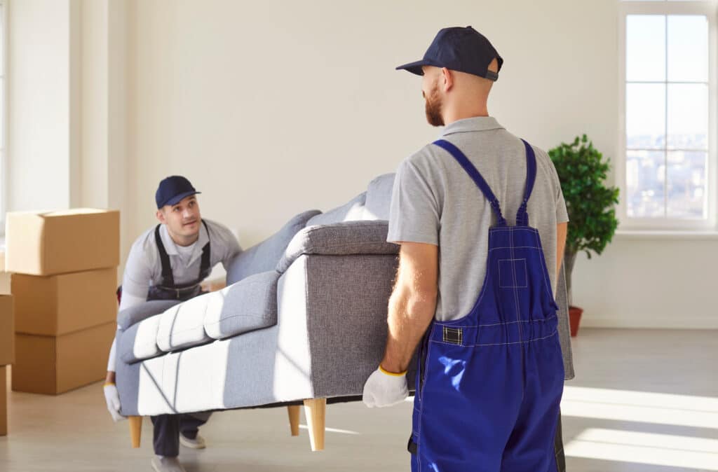 Deux déménageurs professionnels transportant un canapé dans un appartement lumineux avec des cartons empilés en arrière-plan.
